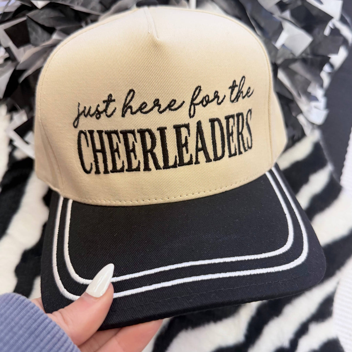 a lightweight unisex embroidered hat with black script 'just here for the' lettering, bold block 'CHEERLEADERS' text, contrasting black bill, white piped trim, cream structured front, black top button, and adjustable snap-back closure