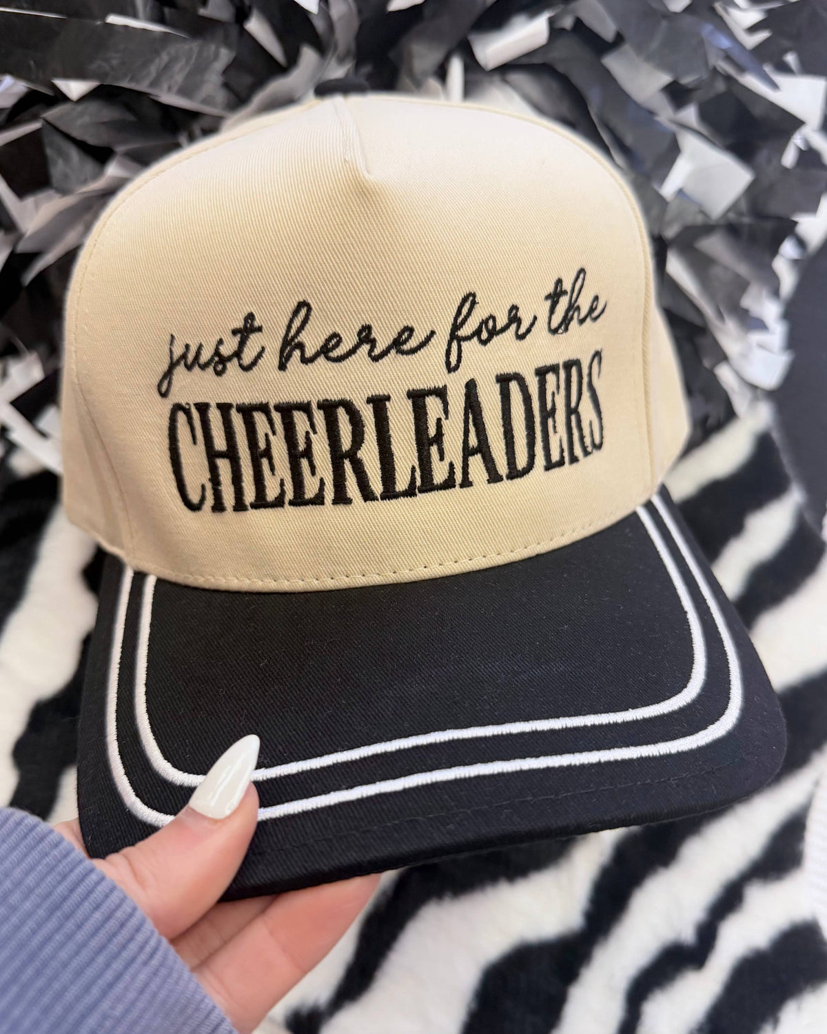 a lightweight unisex embroidered hat with black script 'just here for the' lettering, bold block 'CHEERLEADERS' text, contrasting black bill, white piped trim, cream structured front, black top button, and adjustable snap-back closure