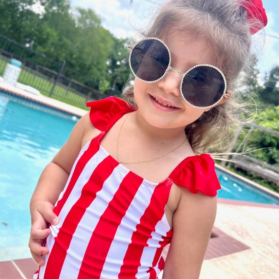 kids' sunglasses with rose gold frame and bridge, black round lenses, rhinestone trim, black temple tips featuring the brand name in white, nose pads, UV protection, and a signature pouch