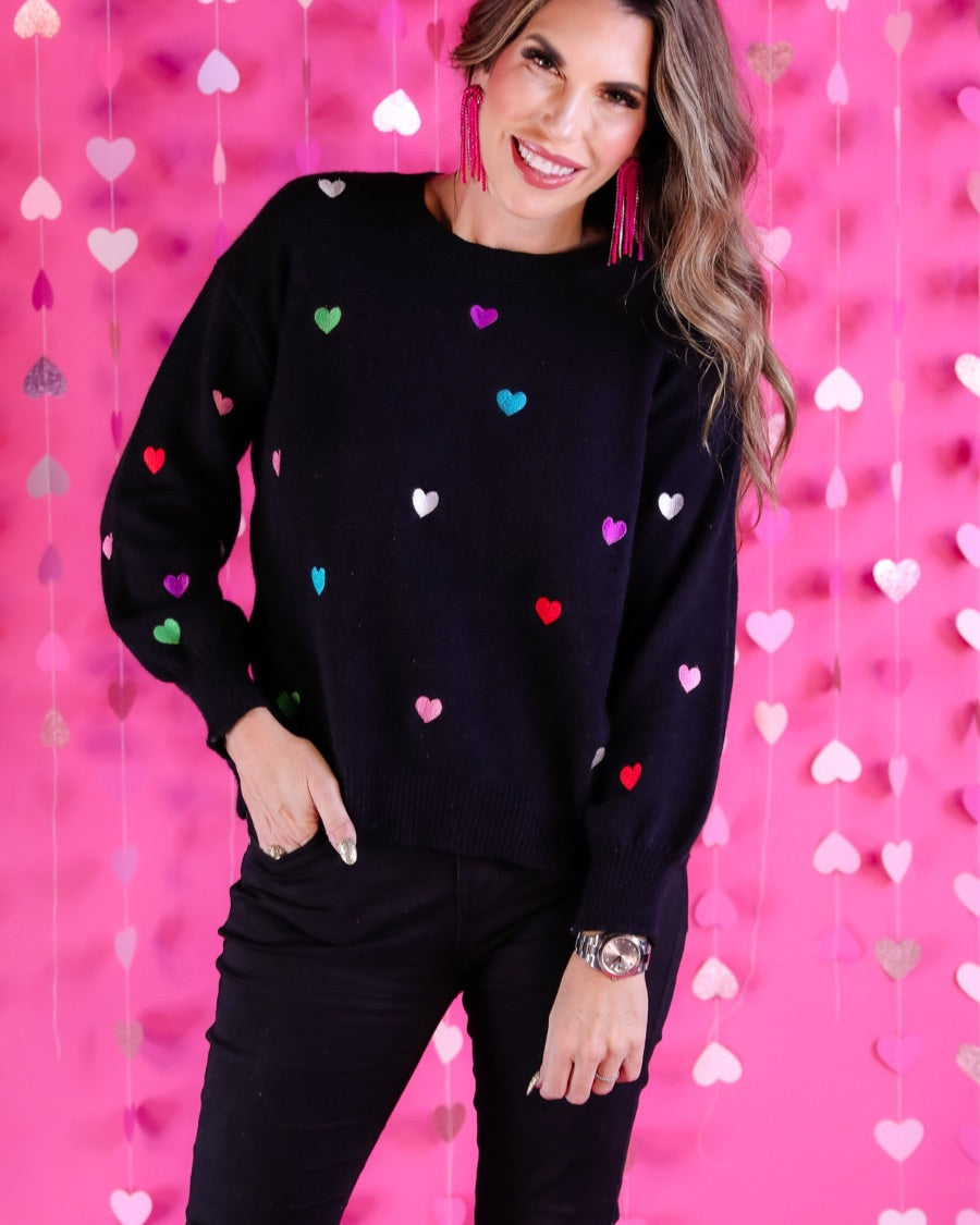 a black long-sleeve sweater crafted from snug, plush material adorned with colorful embroidered hearts, finished with ribbed trim on the neckline, cuffs, and hem
