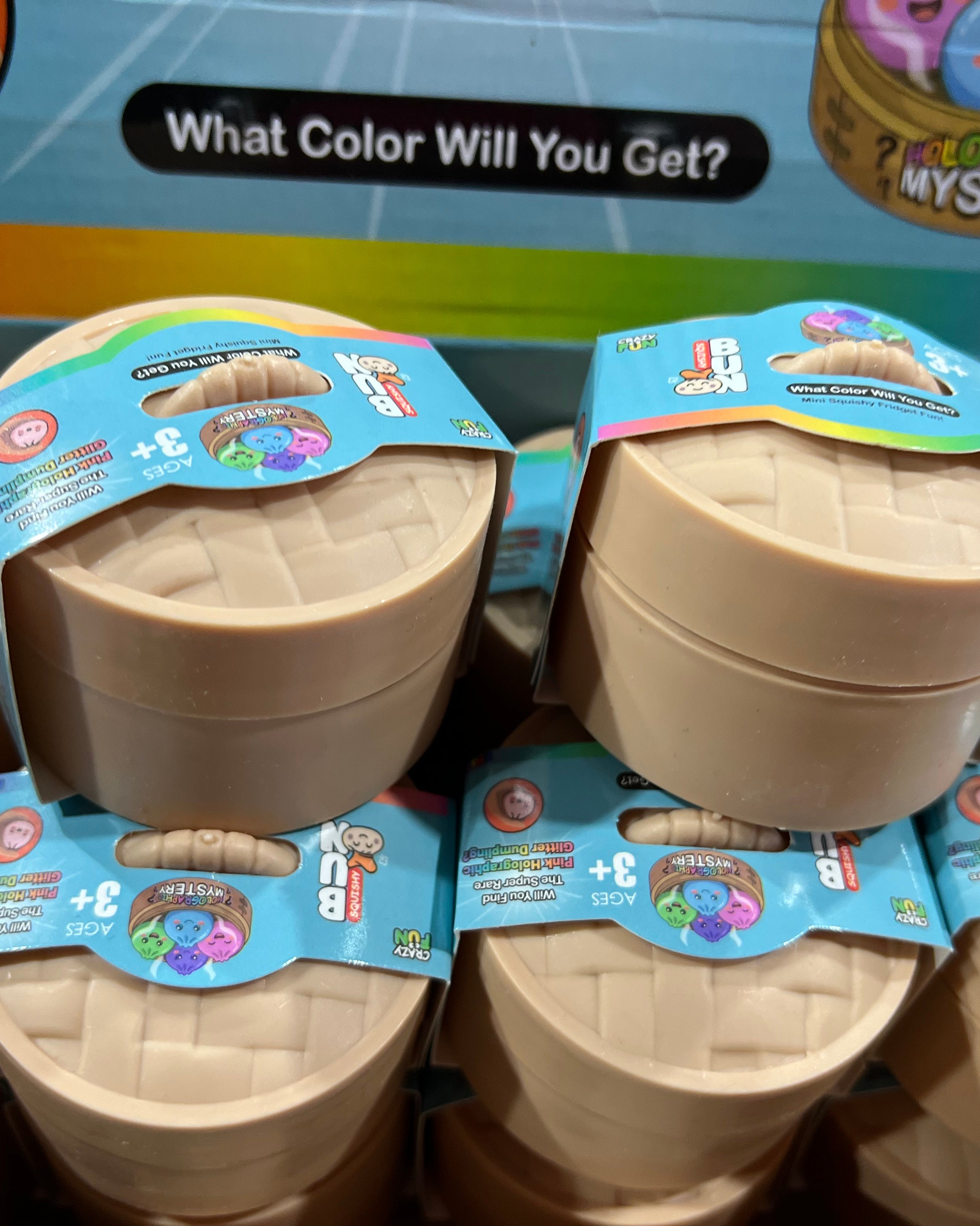 A retail display box filled with small round plastic mystery toy containers. The containers are beige/tan with blue and multicolor labels, and the top box reads 'What Color Will You Get?' and 'Mystery'.