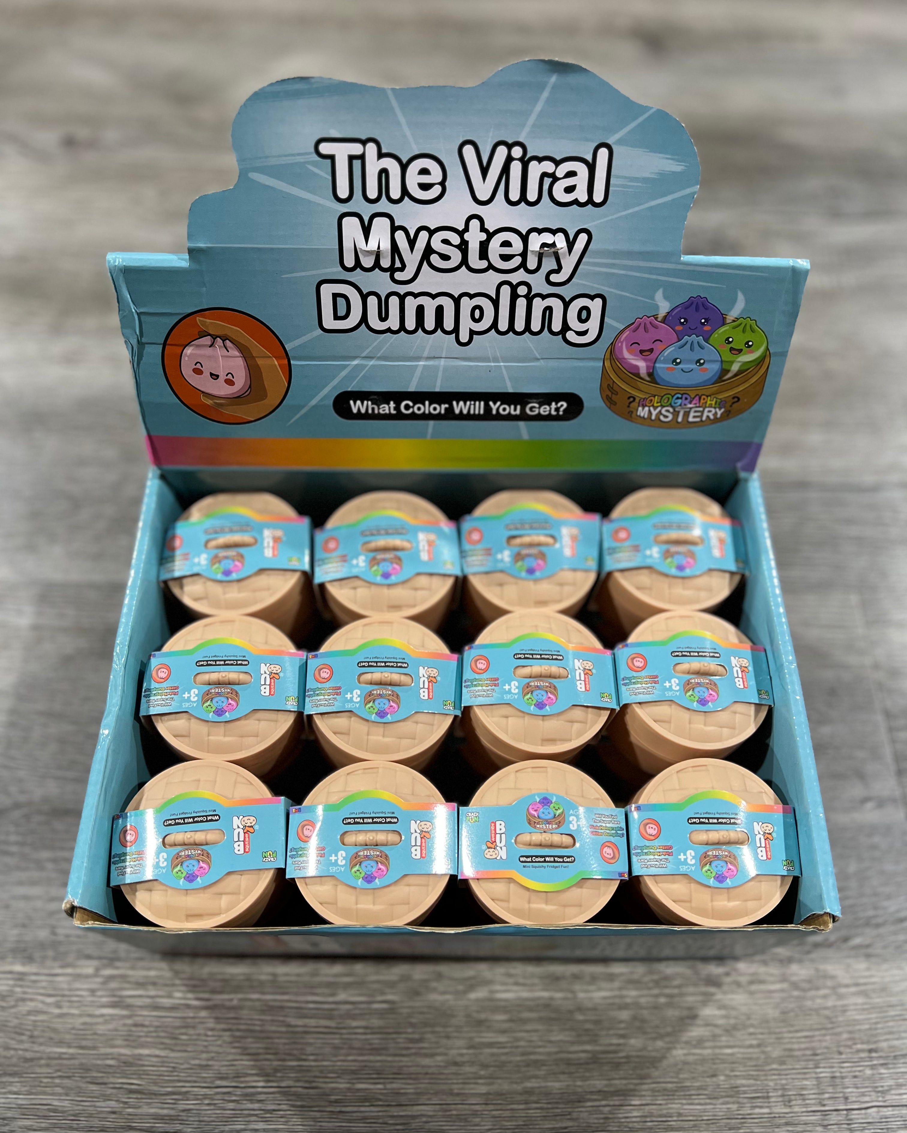 A retail display box of 'The Viral Mystery Dumpling' products, showing twelve individual round packages arranged inside a blue and white cardboard display unit on a gray wood-textured surface.