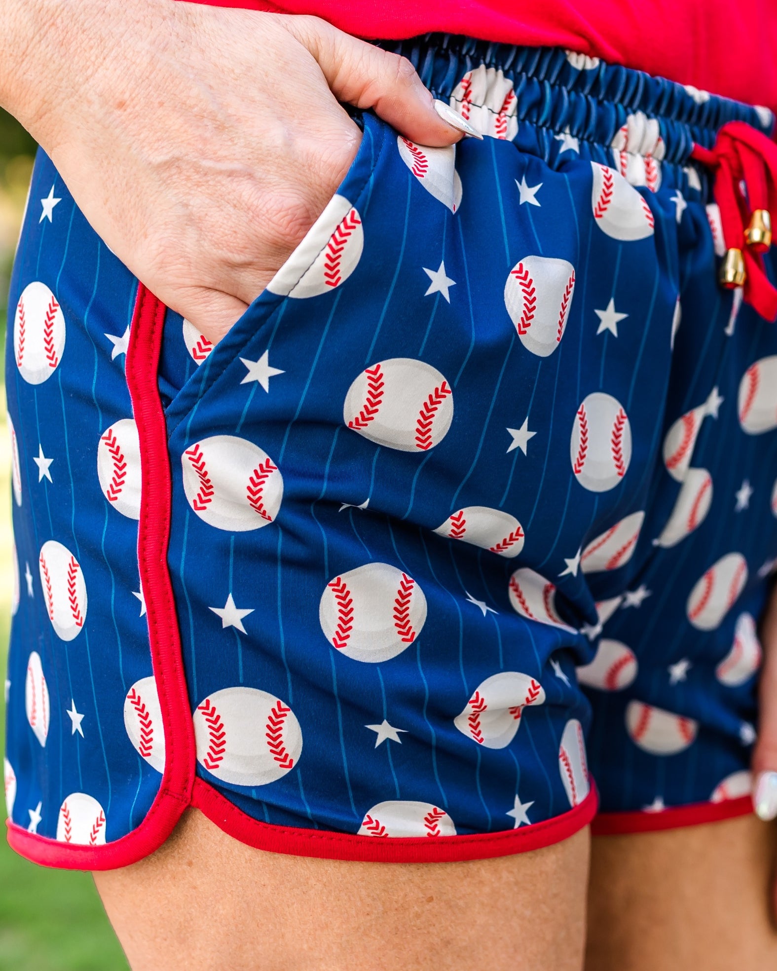 mid-rise relaxed fit activewear shorts with side pockets and a tie waist drawstring with gold bead accents, designed on a baseball pattern print fabric, royal blue base, and red trim