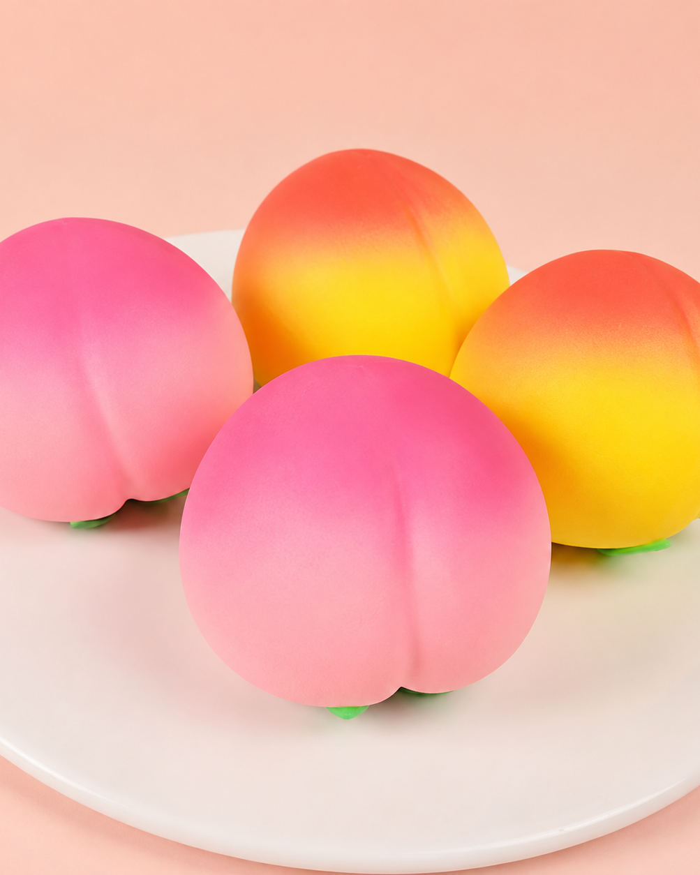 peach-shaped squishy toys with a soft touch texture, rounded fruit design, center groove, green leaf accent, and clear individual packaging in pink ombré and yellow-orange ombré