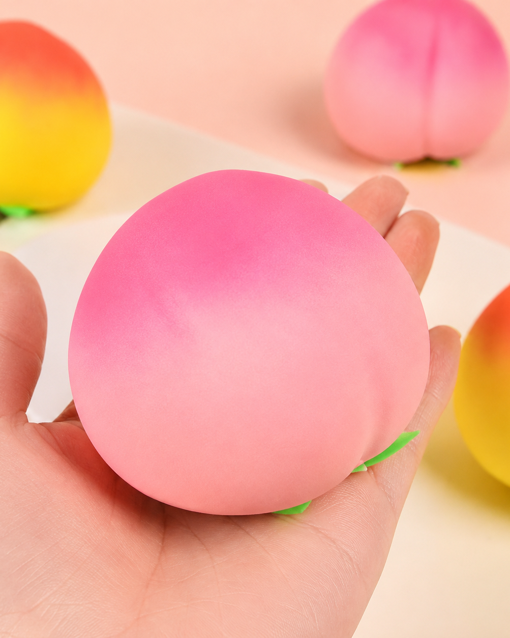 a pink ombré peach-shaped squishy toy with a soft touch texture, rounded fruit design, center groove, green leaf accent, and clear individual packaging