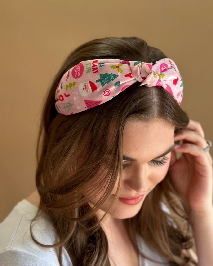 a twisted center top knot headband crafted from soft, lightweight pink fabric with a mix of holiday elements (cupcakes, lollipops, mistletoe, Santa, and trees) and joyful holiday sayings (Fa La La, Joy, Merry Xmas, and Oh What Fun)