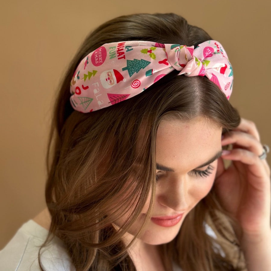 a twisted center top knot headband crafted from soft, lightweight pink fabric with a mix of holiday elements (cupcakes, lollipops, mistletoe, Santa, and trees) and joyful holiday sayings (Fa La La, Joy, Merry Xmas, and Oh What Fun)