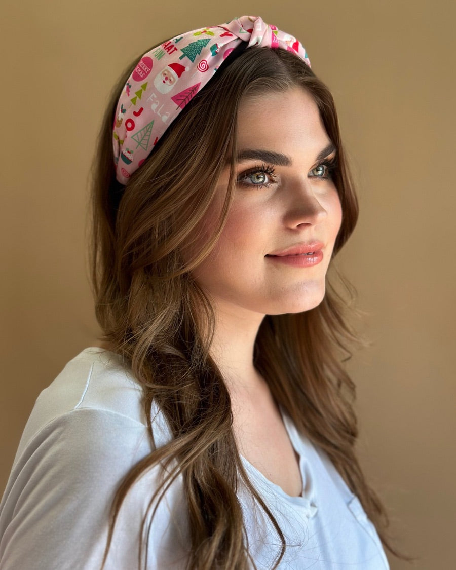 a twisted center top knot headband crafted from soft, lightweight pink fabric with a mix of holiday elements (cupcakes, lollipops, mistletoe, Santa, and trees) and joyful holiday sayings (Fa La La, Joy, Merry Xmas, and Oh What Fun)