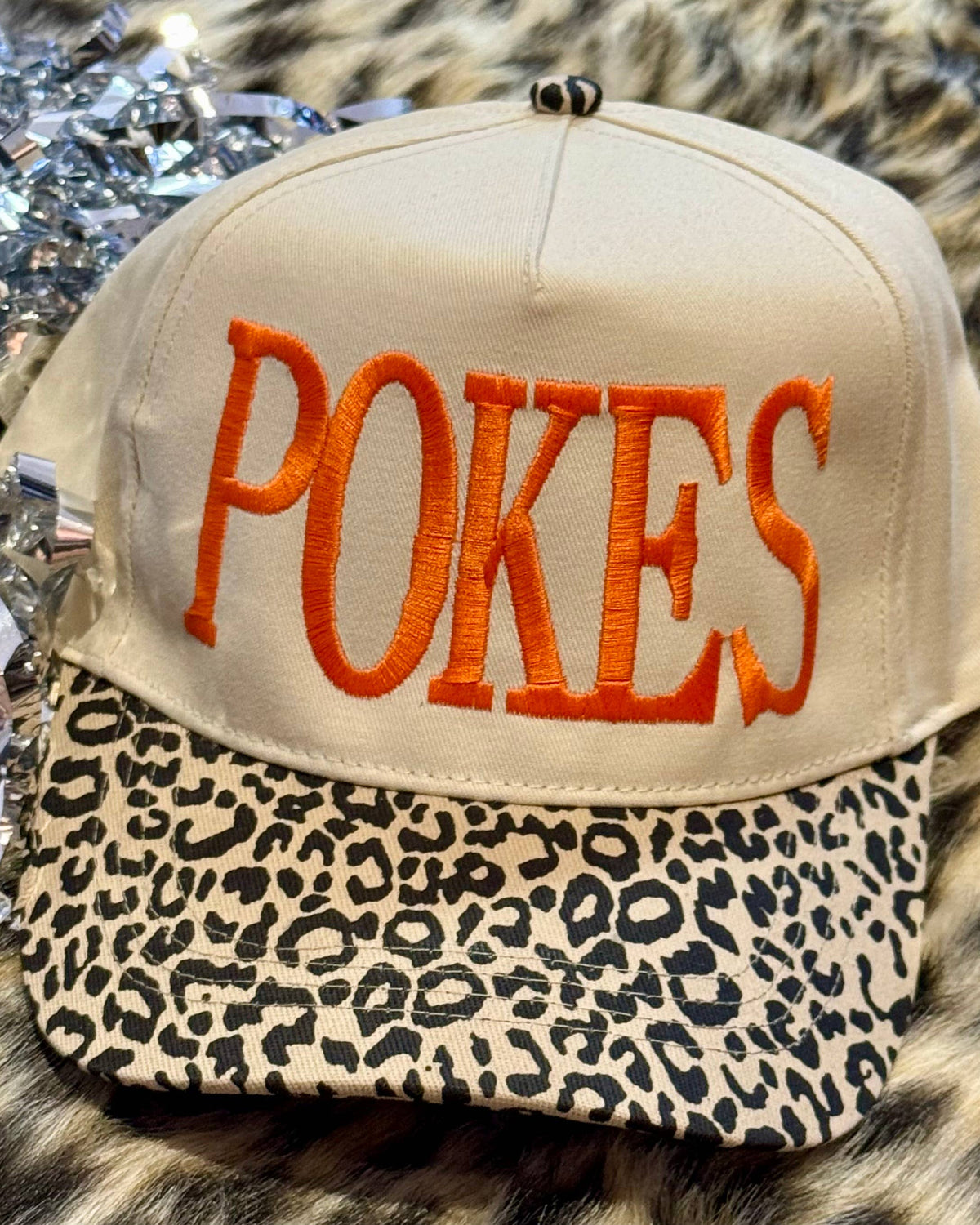 a lightweight unisex two-tone hat with embroidered orange 'POKES' lettering, cream structured front, curved contrasting black leopard bill, coordinating leopard top button, and adjustable snap-back closure