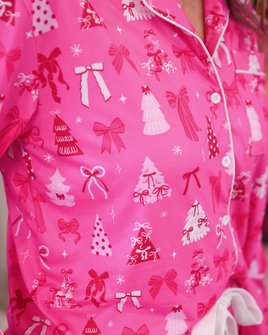 A close-up of a bright pink pajama top with a white and red Christmas print featuring bows, trees, and gifts. A white tie belt is visible at the waist.