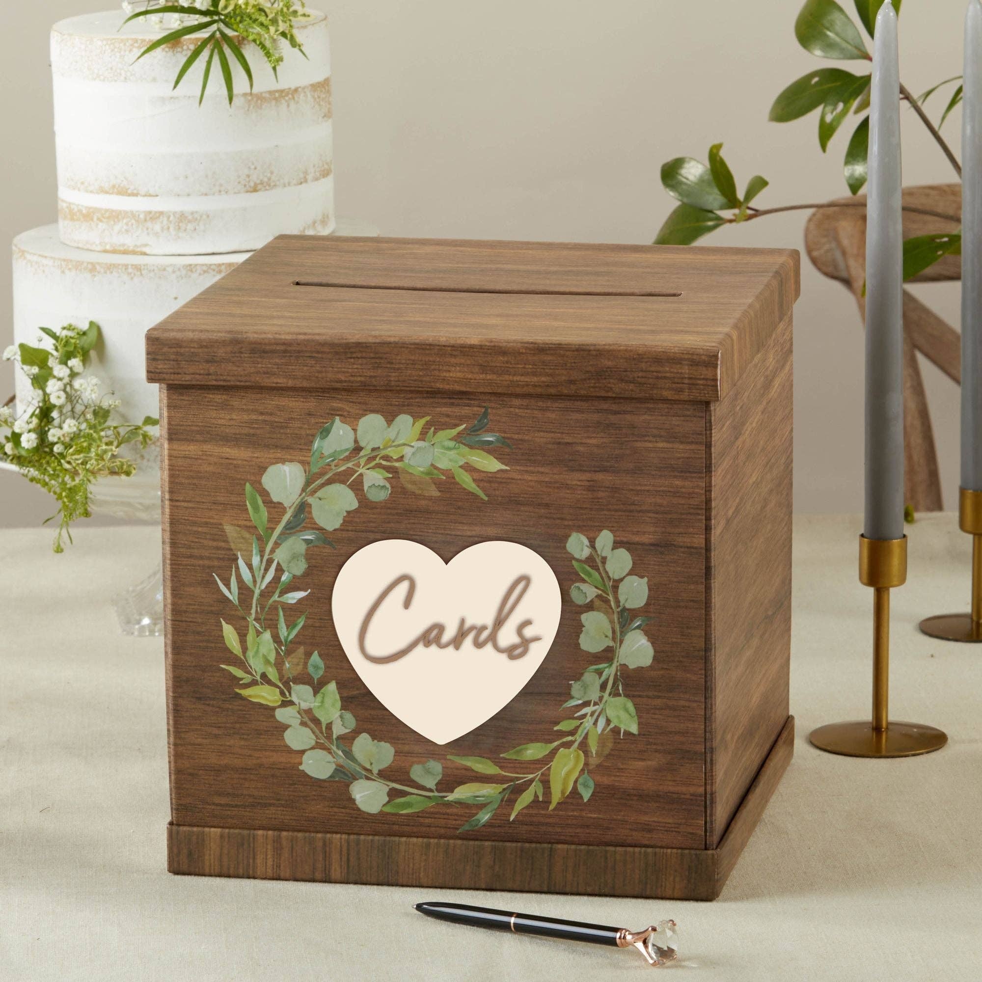 Wooden card box with decorative heart and foliage design, placed on a table with a cake and flowers in the background.