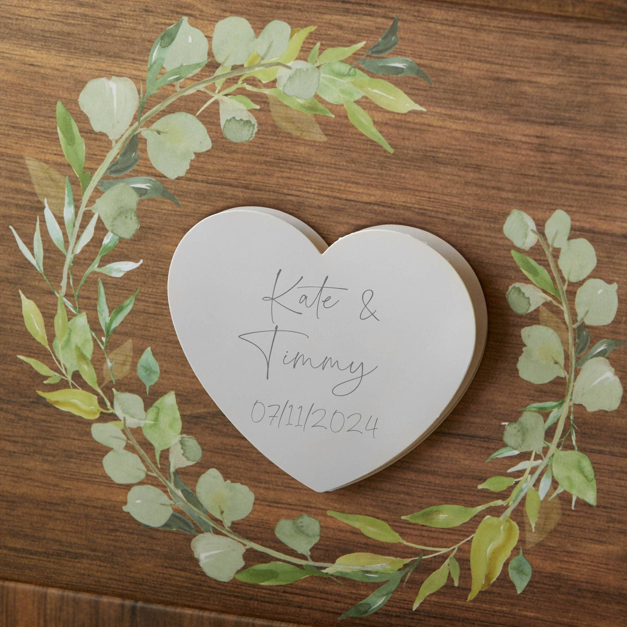 Heart-shaped card with engraved names and date on a wooden background with greenery