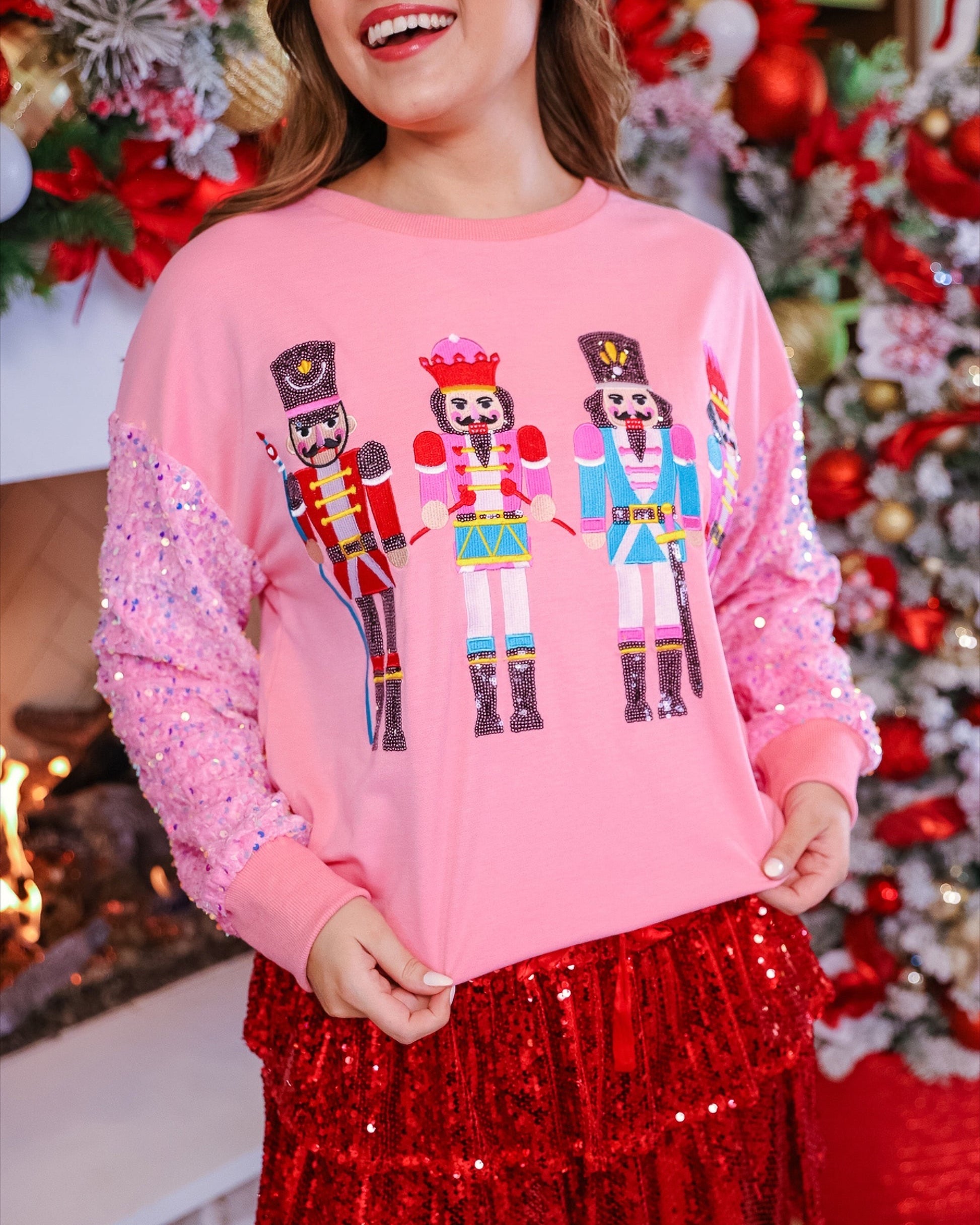 a lightweight pink long-sleeve top decorated with four vibrant nutcracker designs finished with dimensional beaded effect boots and accented by soft, shimmering velvet-sequin sleeves, a relaxed drop shoulder, and ribbed crew neckline, cuffs, and hem