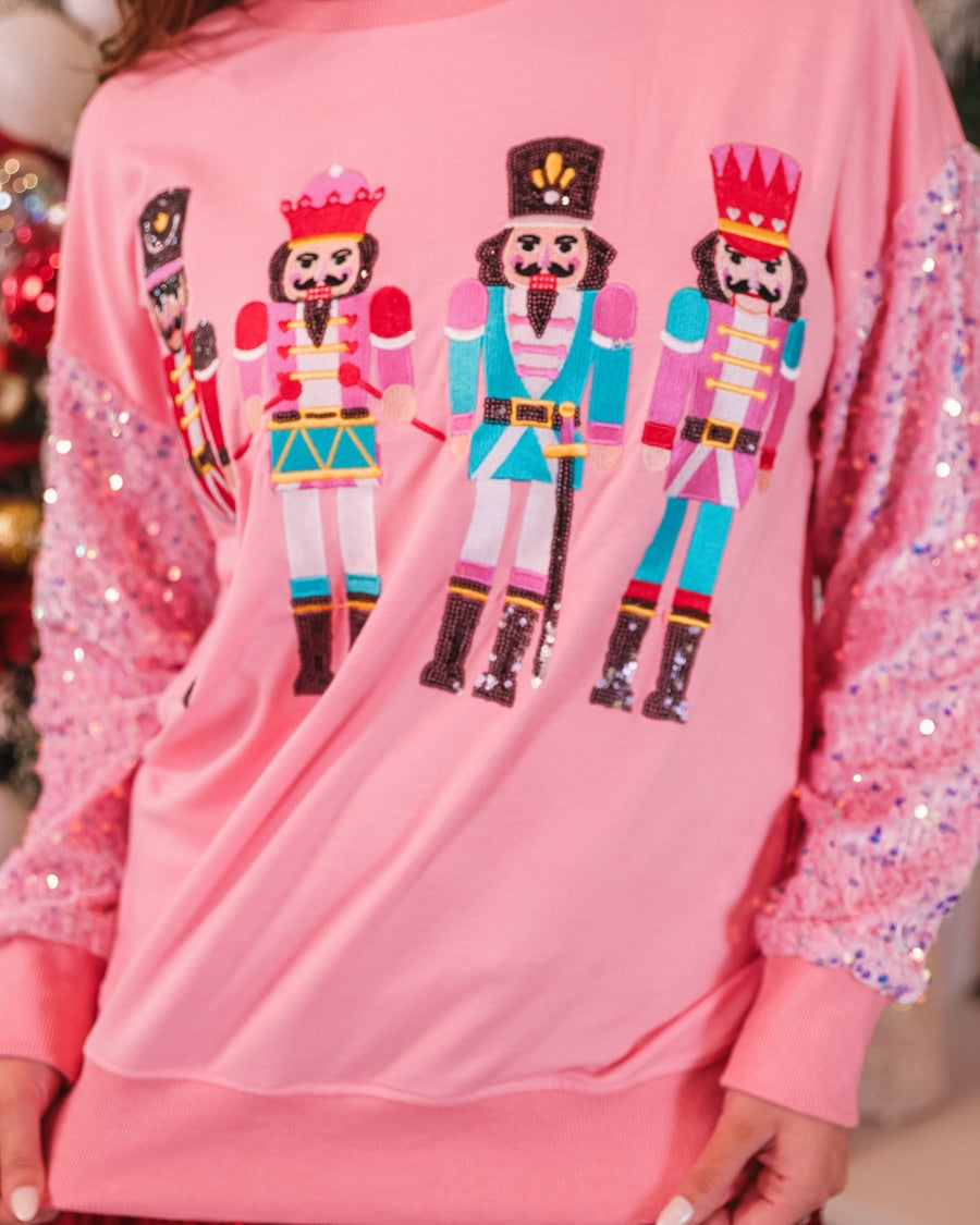 a lightweight pink long-sleeve top decorated with four vibrant nutcracker designs finished with dimensional beaded effect boots and accented by soft, shimmering velvet-sequin sleeves, a relaxed drop shoulder, and ribbed crew neckline, cuffs, and hem