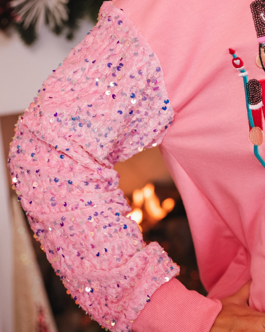 a lightweight pink long-sleeve top decorated with four vibrant nutcracker designs finished with dimensional beaded effect boots and accented by soft, shimmering velvet-sequin sleeves, a relaxed drop shoulder, and ribbed crew neckline, cuffs, and hem