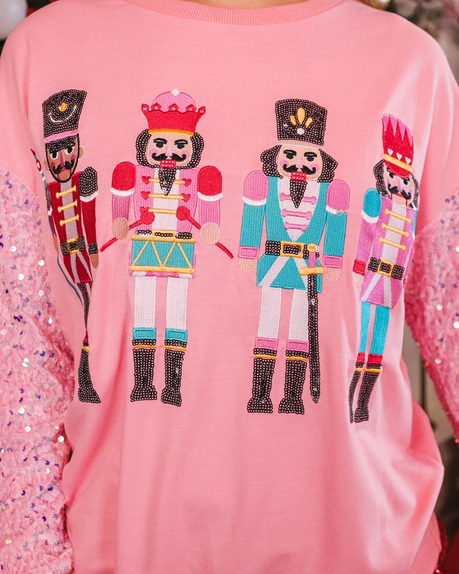 a lightweight pink long-sleeve top decorated with four vibrant nutcracker designs finished with dimensional beaded effect boots and accented by soft, shimmering velvet-sequin sleeves, a relaxed drop shoulder, and ribbed crew neckline, cuffs, and hem