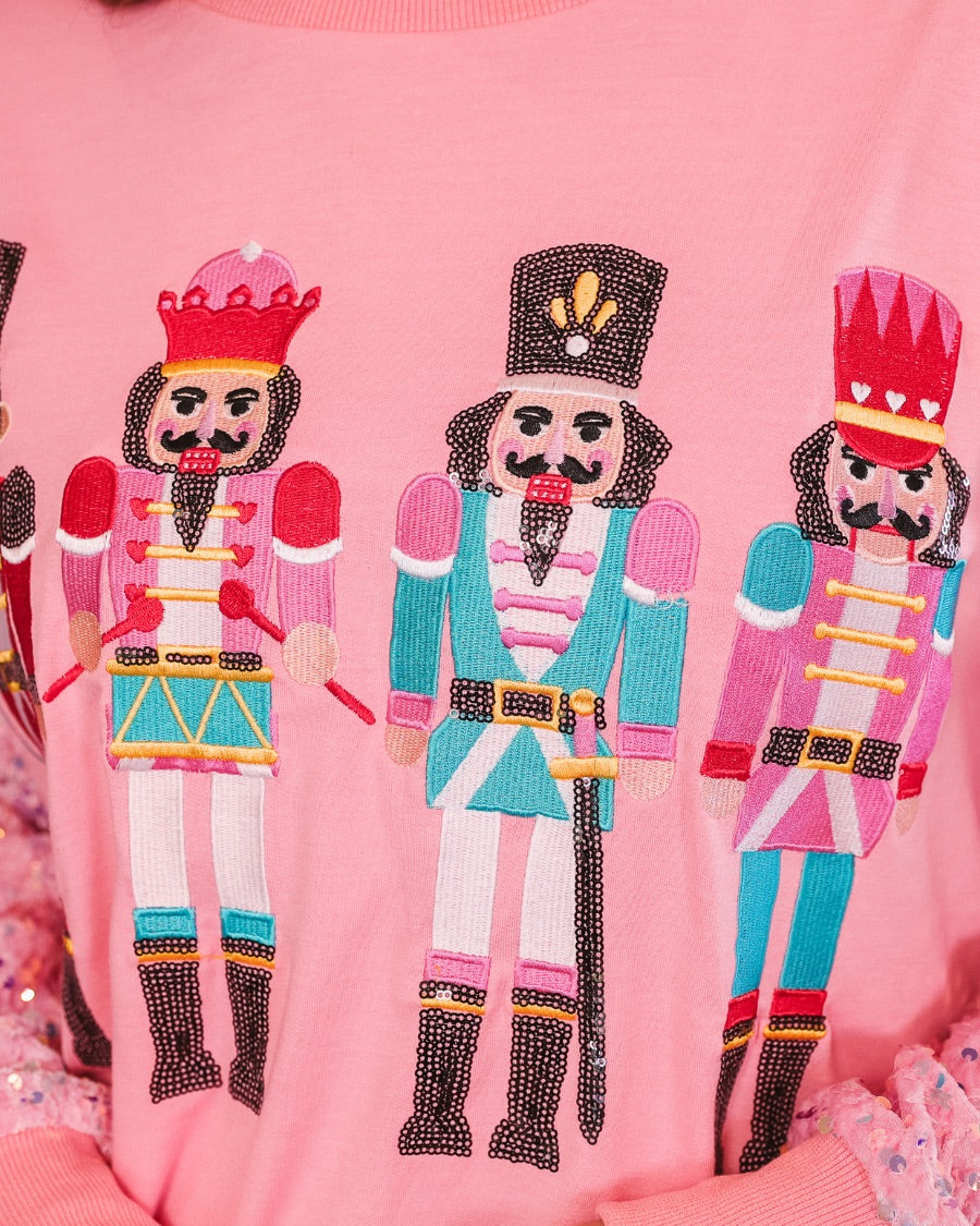 a lightweight pink long-sleeve top decorated with four vibrant nutcracker designs finished with dimensional beaded effect boots and accented by soft, shimmering velvet-sequin sleeves, a relaxed drop shoulder, and ribbed crew neckline, cuffs, and hem