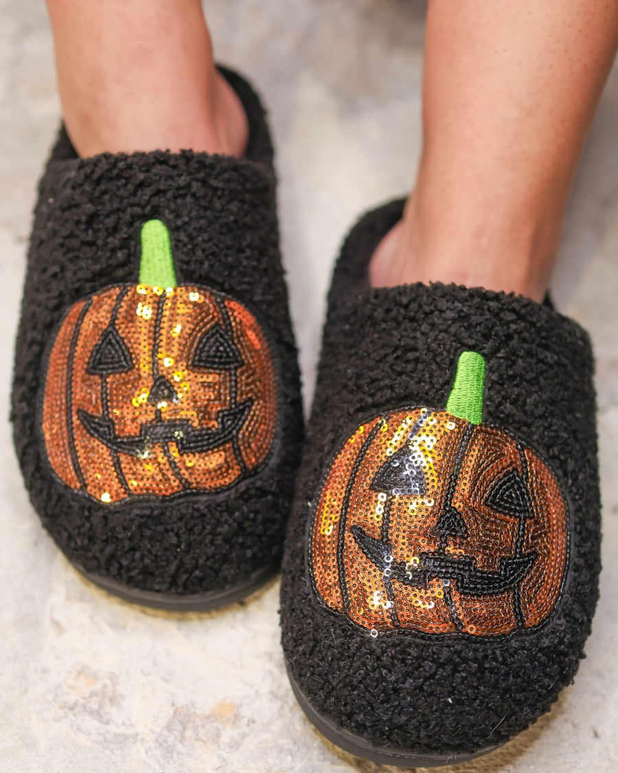 black fuzzy mule slippers with orange sequin jack-o'-lantern appliqués, embroidered green stem accents, cushioned flatbeds, plush lining, open back design, and textured bottoms