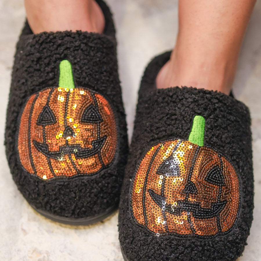 black fuzzy mule slippers with orange sequin jack-o'-lantern appliqués, embroidered green stem accents, cushioned flatbeds, plush lining, open back design, and textured bottoms