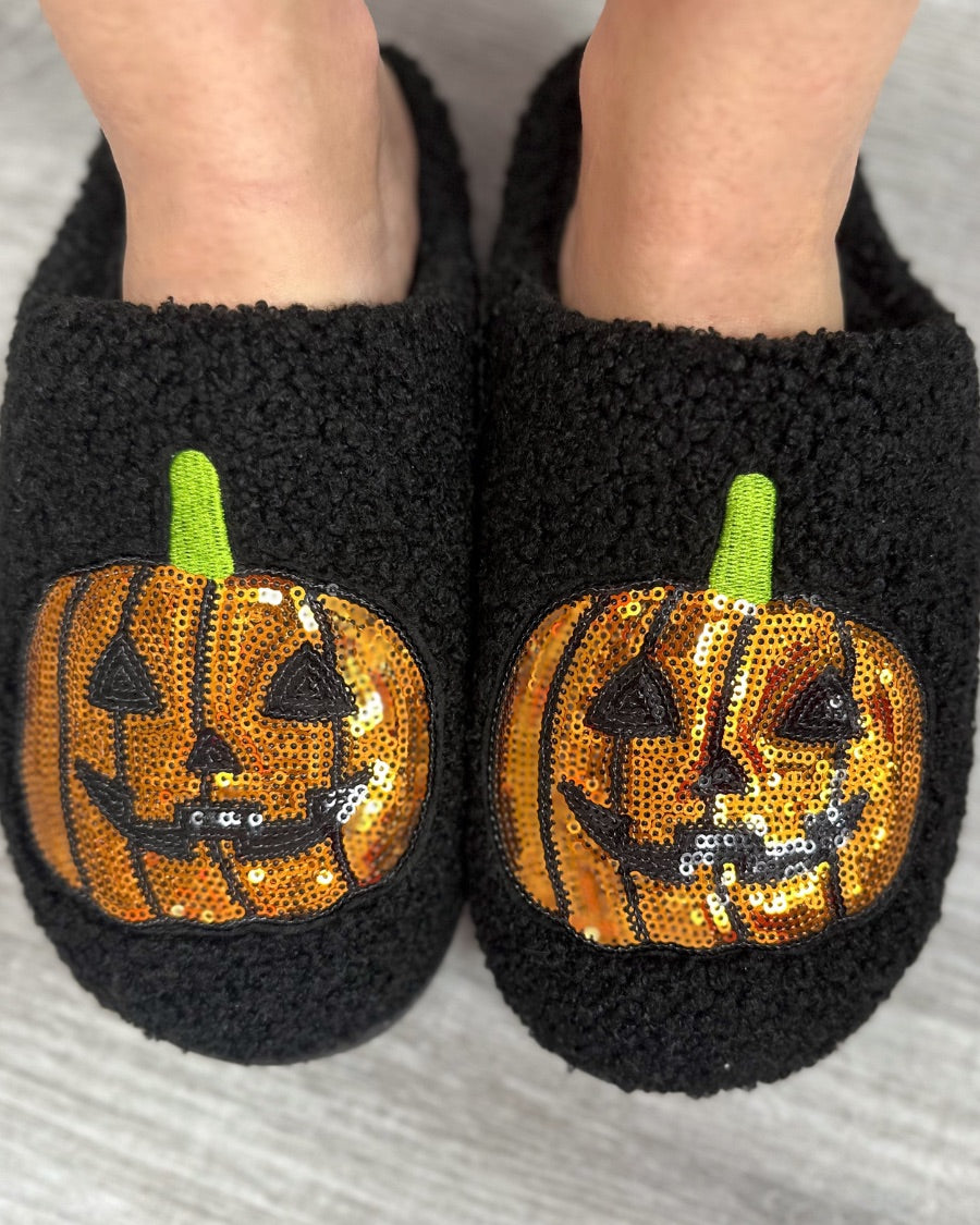 black fuzzy mule slippers with orange sequin jack-o'-lantern appliqués, embroidered green stem accents, cushioned flatbeds, plush lining, open back design, and textured bottoms