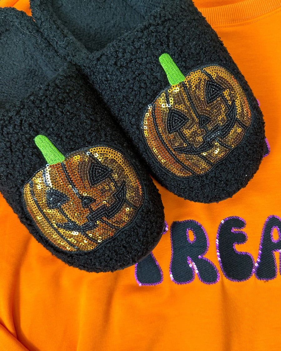 black fuzzy mule slippers with orange sequin jack-o'-lantern appliqués, embroidered green stem accents, cushioned flatbeds, plush lining, open back design, and textured bottoms