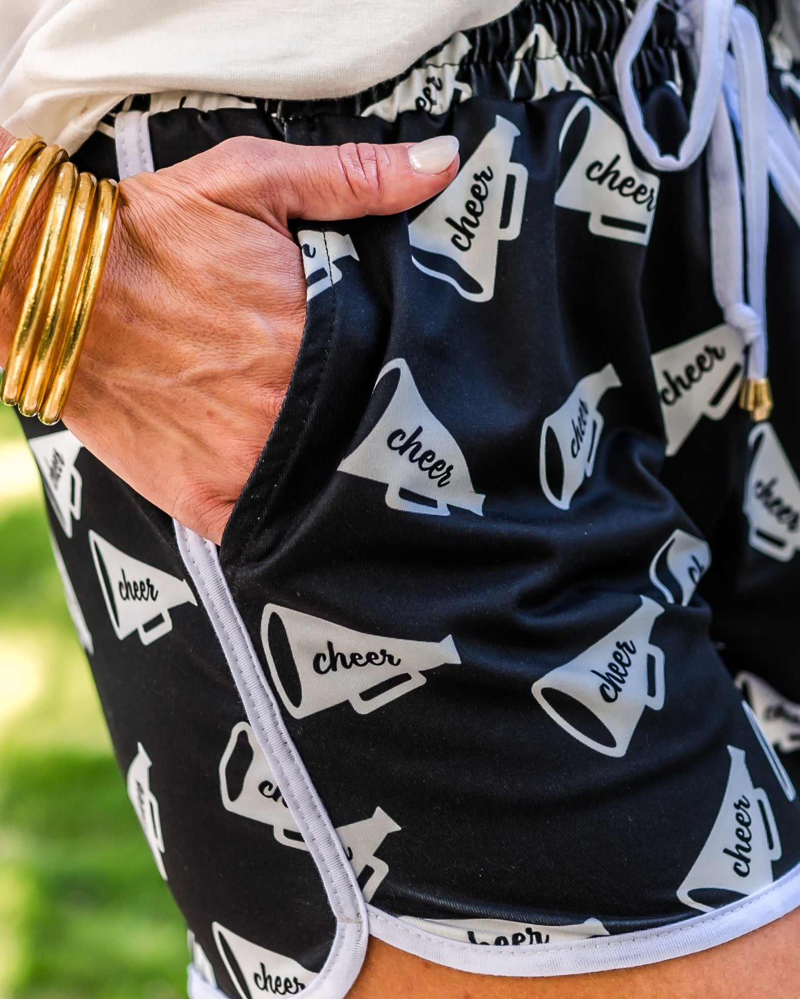 mid-rise relaxed fit activewear shorts with side pockets and a tie waist drawstring with gold bead accents, designed on a white megaphone with the word "cheer" handwritten across the center pattern print fabric, black base, and white trim