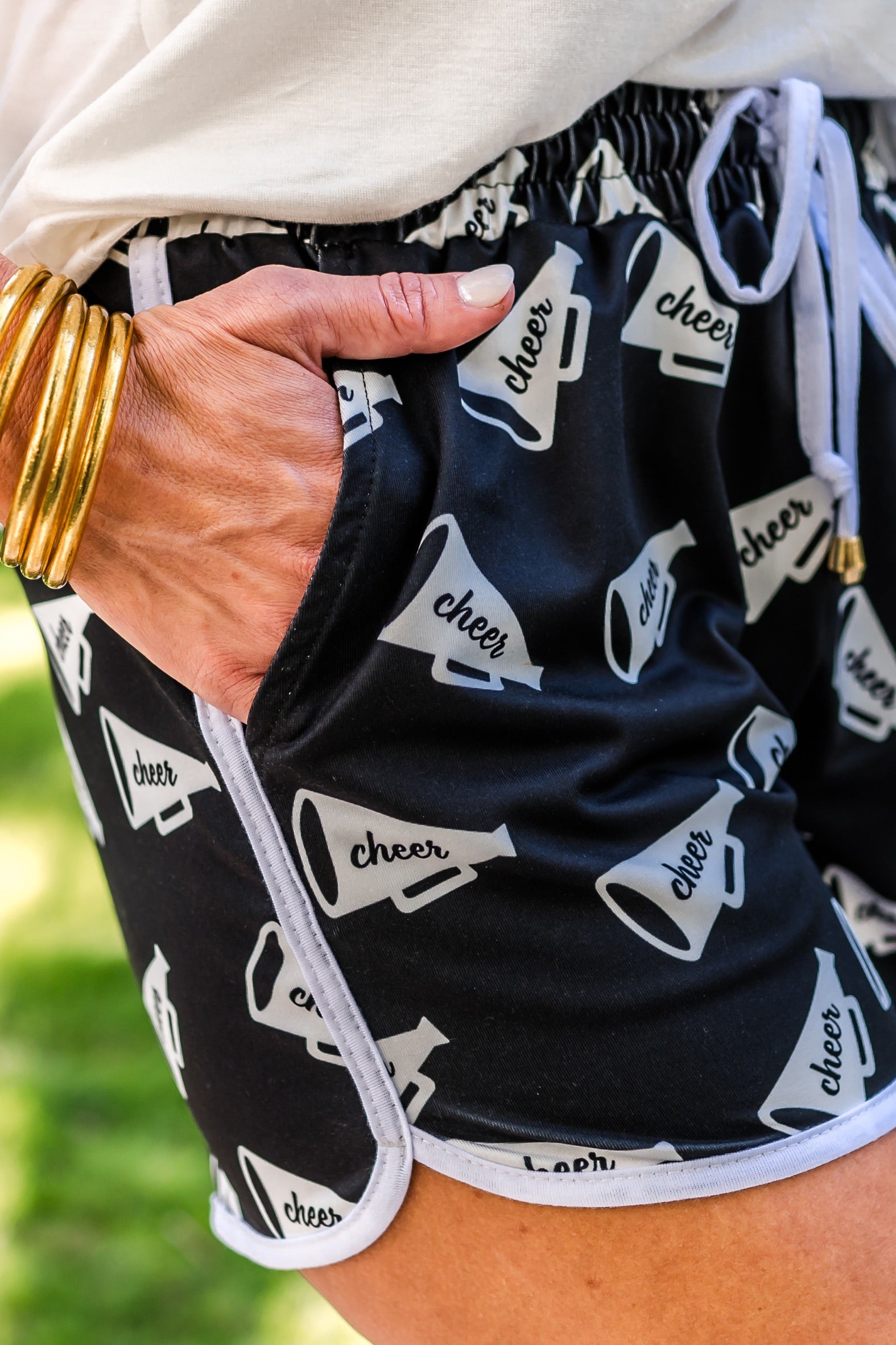 mid-rise relaxed fit activewear shorts with side pockets and a tie waist drawstring with gold bead accents, designed on a white megaphone with the word "cheer" handwritten across the center pattern print fabric, black base, and white trim