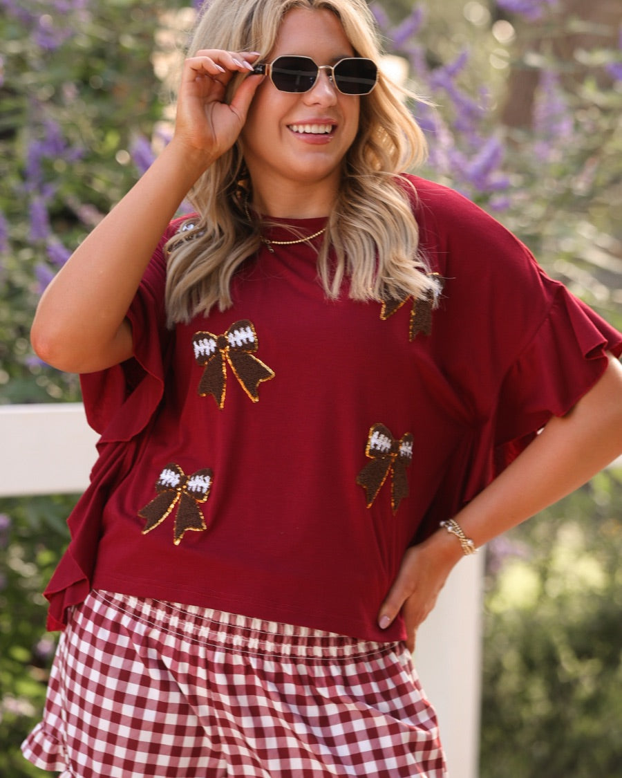 fluttery ruffle short-sleeve top with a mom crop length, sewn-on chenille football bow patches outlined in sequins on a soft, stretch-knit fabric in maroon