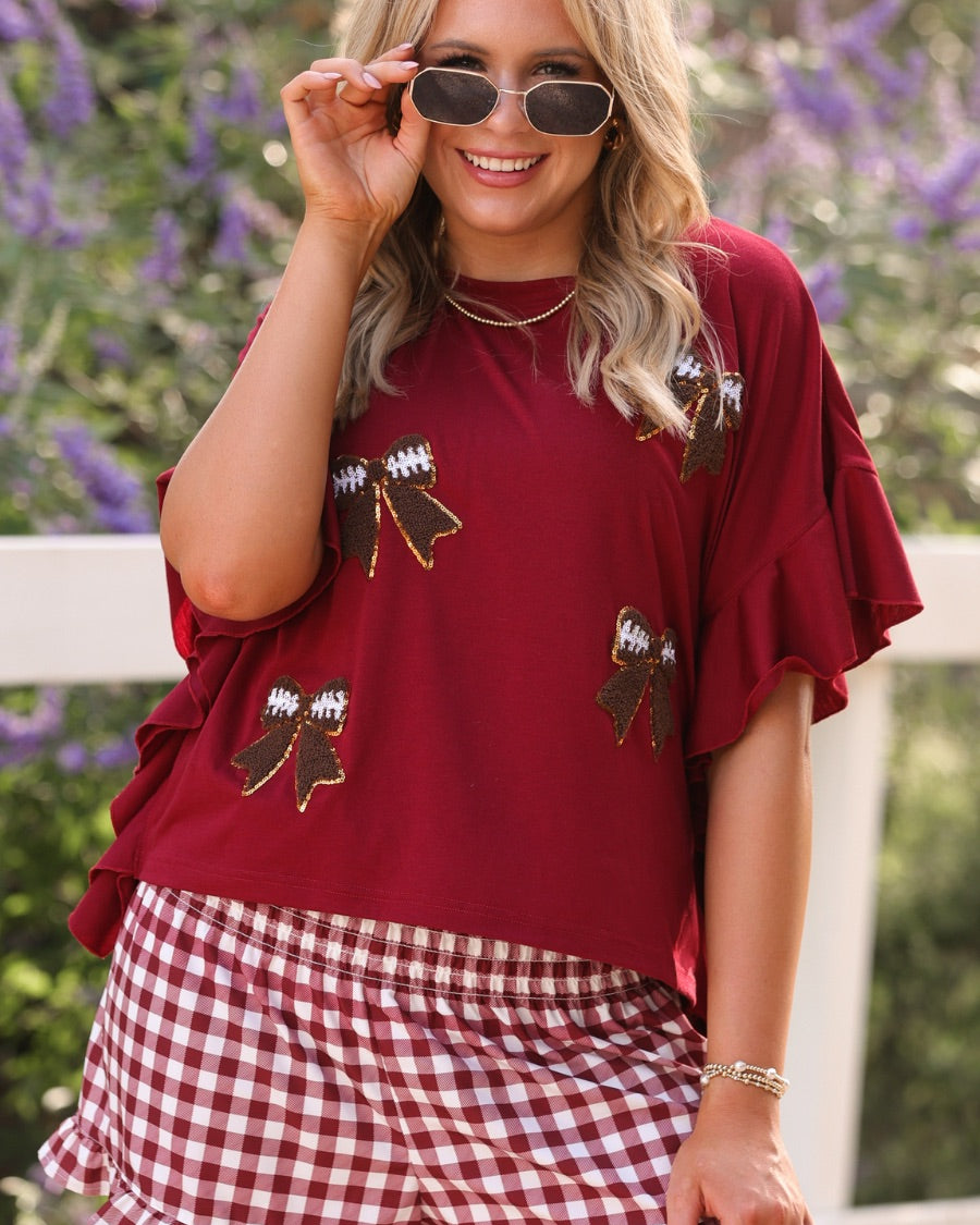 fluttery ruffle short-sleeve top with a mom crop length, sewn-on chenille football bow patches outlined in sequins on a soft, stretch-knit fabric in maroon