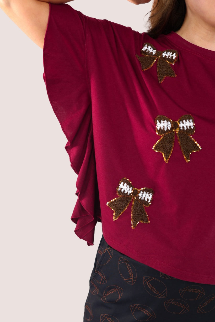 fluttery ruffle short-sleeve top with a mom crop length, sewn-on chenille football bow patches outlined in sequins on a soft, stretch-knit fabric in maroon