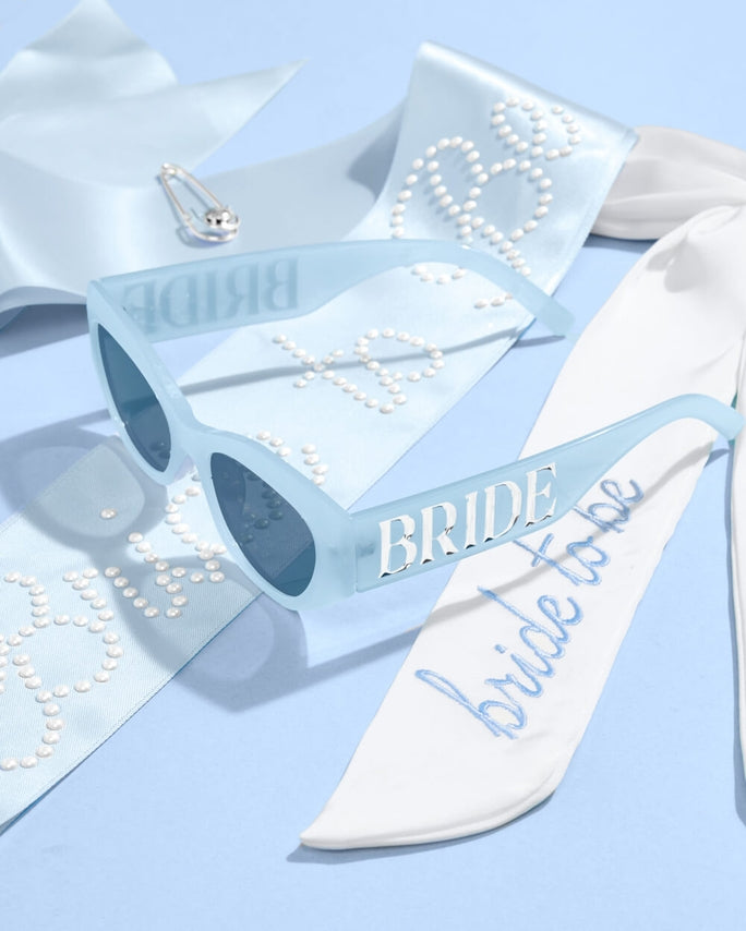 Blue sunglasses and 'Bride to be' sash on a light blue background