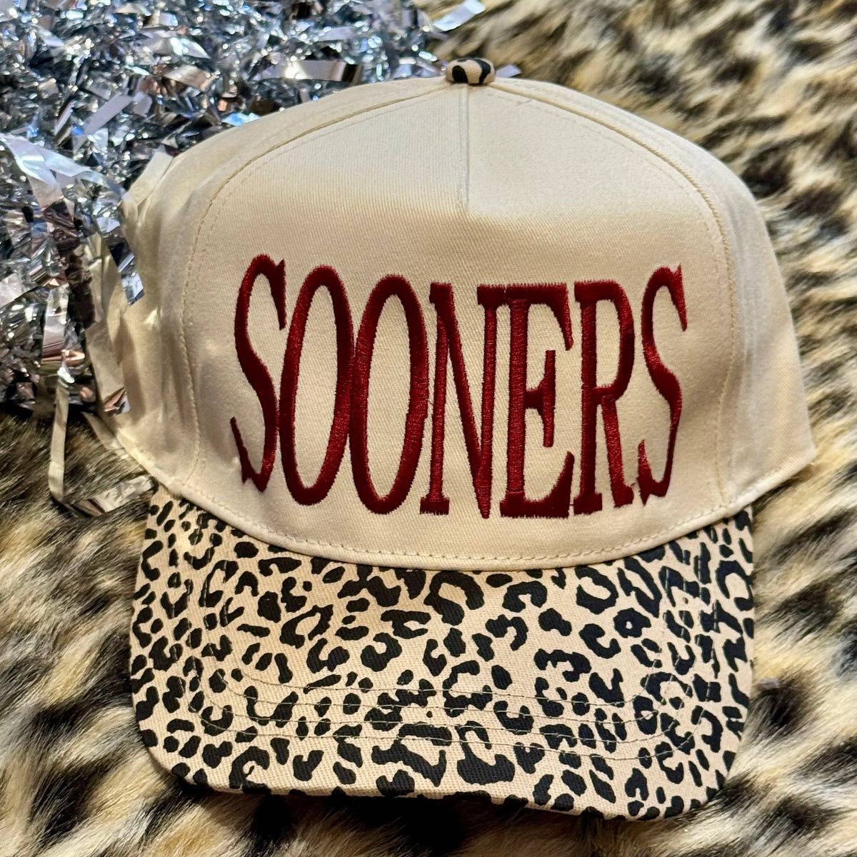 a lightweight unisex two-tone hat with embroidered crimson 'SOONERS' lettering, cream structured front, curved contrasting black leopard bill, coordinating leopard top button, and adjustable snap-back closure