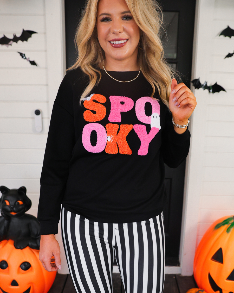 a black long-sleeve pullover made of ultra soft luxe material with bold plush chenille 'SPOOKY' patch lettering (orange and pink), white embroidered character accents (tiny bat, ghost and spider), ribbed cuffs, a banded hem, and a relaxed fit for a classic silhouette
