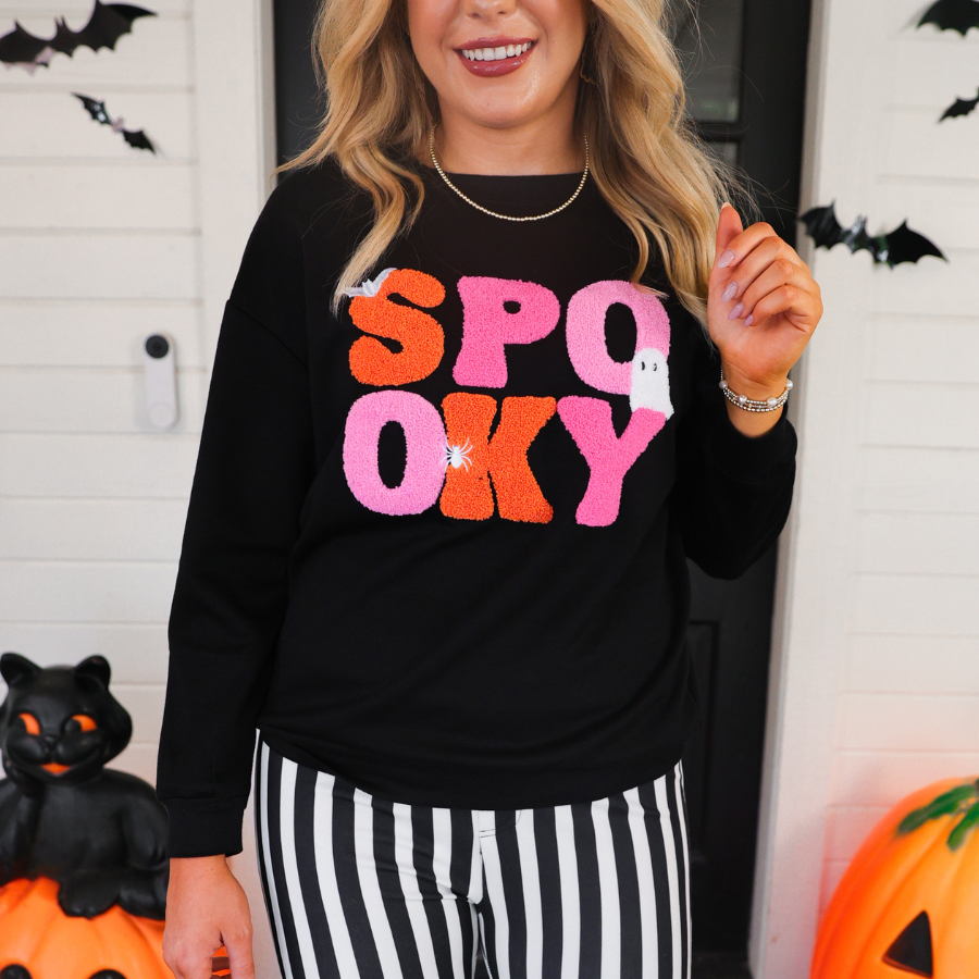 a black long-sleeve pullover made of ultra soft luxe material with bold plush chenille 'SPOOKY' patch lettering (orange and pink), white embroidered character accents (tiny bat, ghost and spider), ribbed cuffs, a banded hem, and a relaxed fit for a classic silhouette