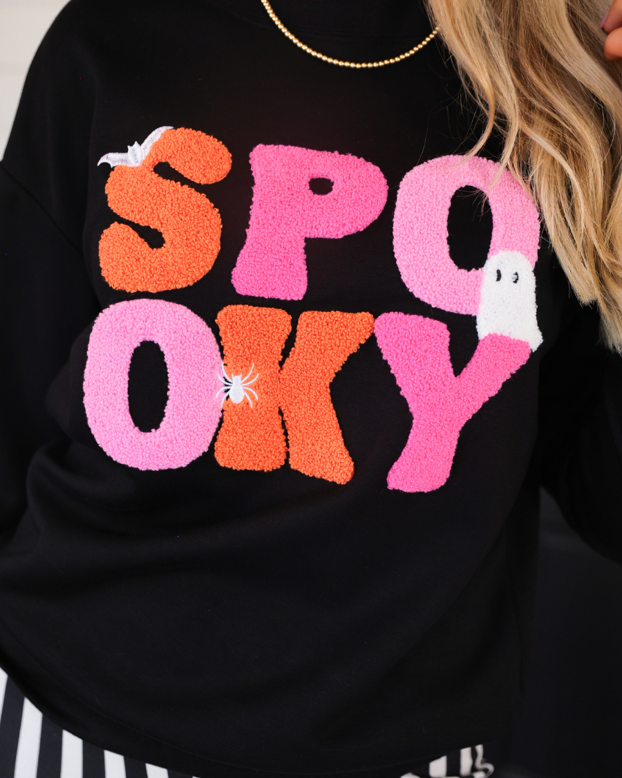 a black long-sleeve pullover made of ultra soft luxe material with bold plush chenille 'SPOOKY' patch lettering (orange and pink), white embroidered character accents (tiny bat, ghost and spider), ribbed cuffs, a banded hem, and a relaxed fit for a classic silhouette