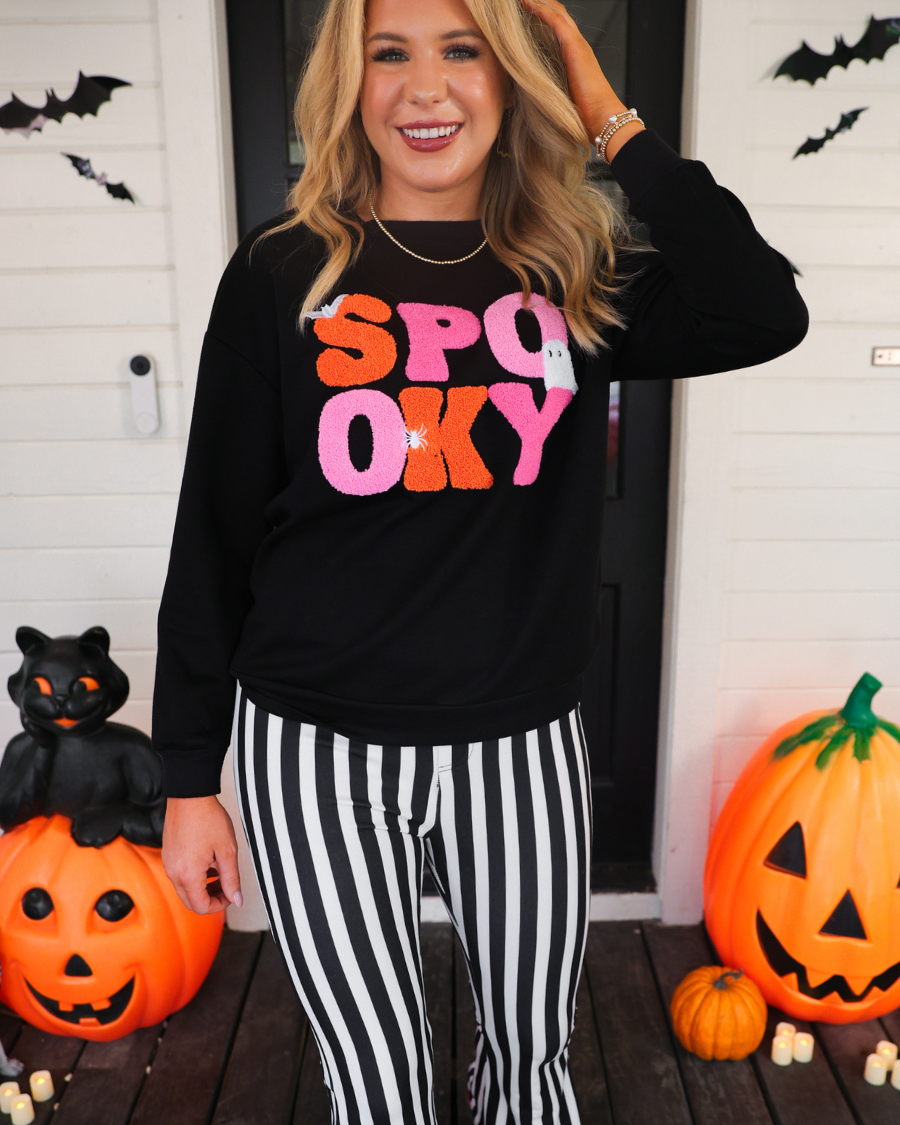 a black long-sleeve pullover made of ultra soft luxe material with bold plush chenille 'SPOOKY' patch lettering (orange and pink), white embroidered character accents (tiny bat, ghost and spider), ribbed cuffs, a banded hem, and a relaxed fit for a classic silhouette