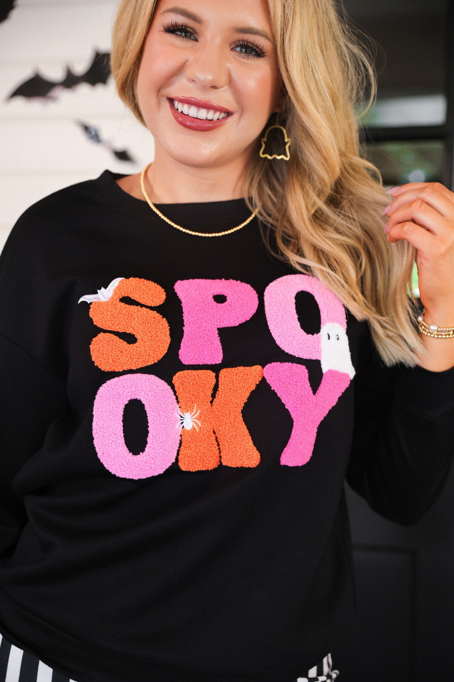 a black long-sleeve pullover made of ultra soft luxe material with bold plush chenille 'SPOOKY' patch lettering (orange and pink), white embroidered character accents (tiny bat, ghost and spider), ribbed cuffs, a banded hem, and a relaxed fit for a classic silhouette
