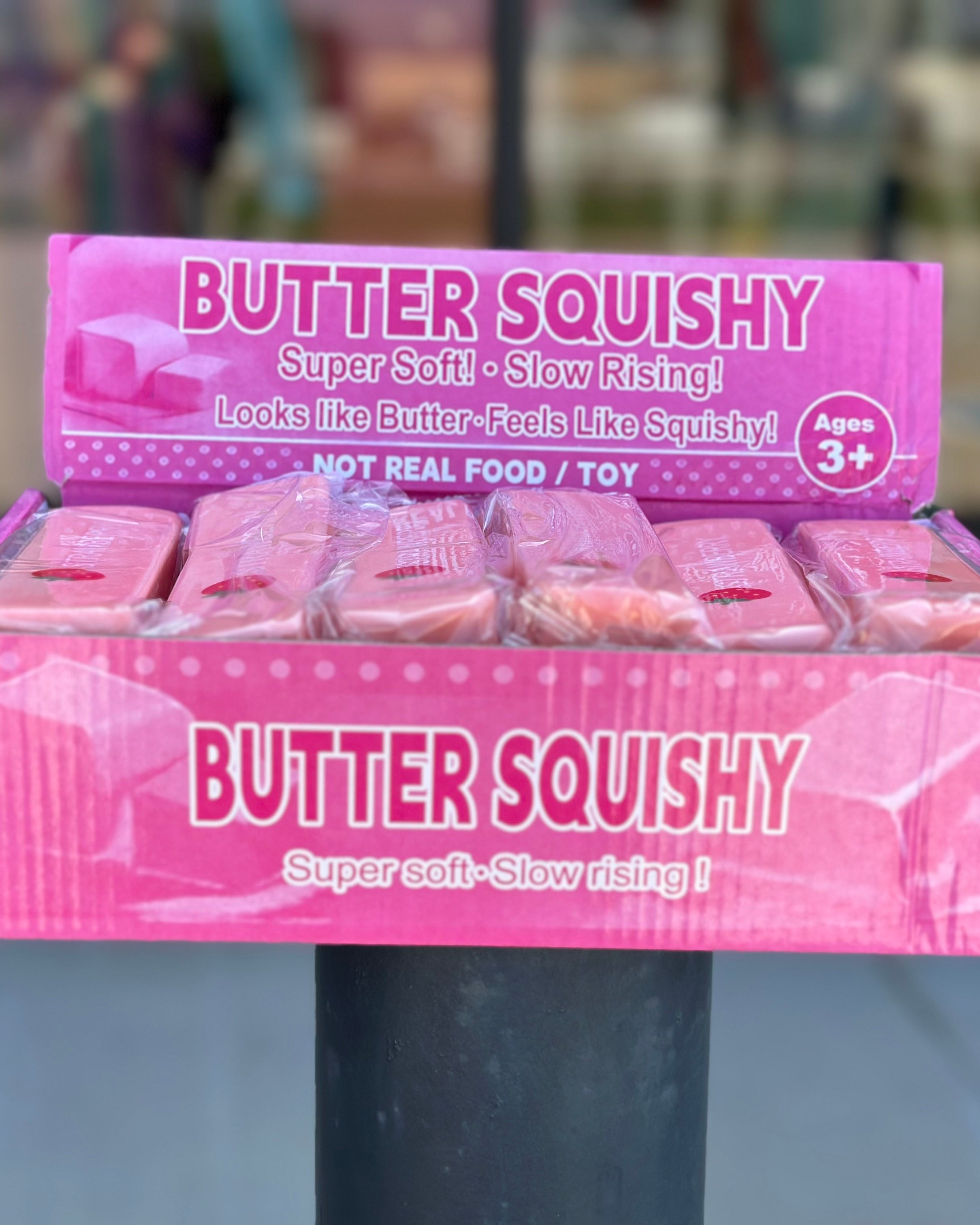 A display box of Butter Squishy toys on a black post outdoors. The box is pink with white text reading 'BUTTER SQUISHY' and shows several individually wrapped pink squishy toys inside.
