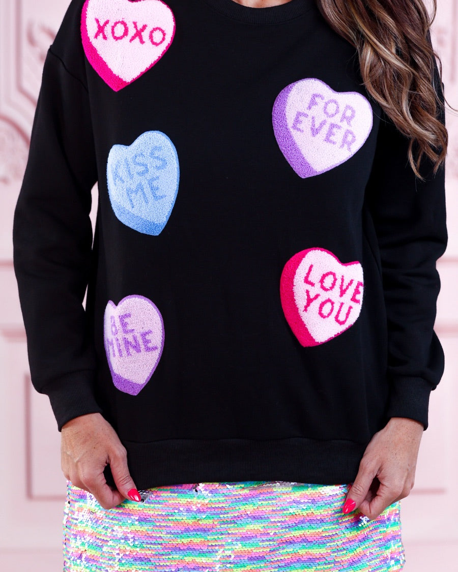 a black long-sleeve sweater made of ultra-soft material adorned with multicolor chenille patch conversation hearts, a relaxed drop-shoulder silhouette, and ribbed trim at the neckline, cuffs, and hemline