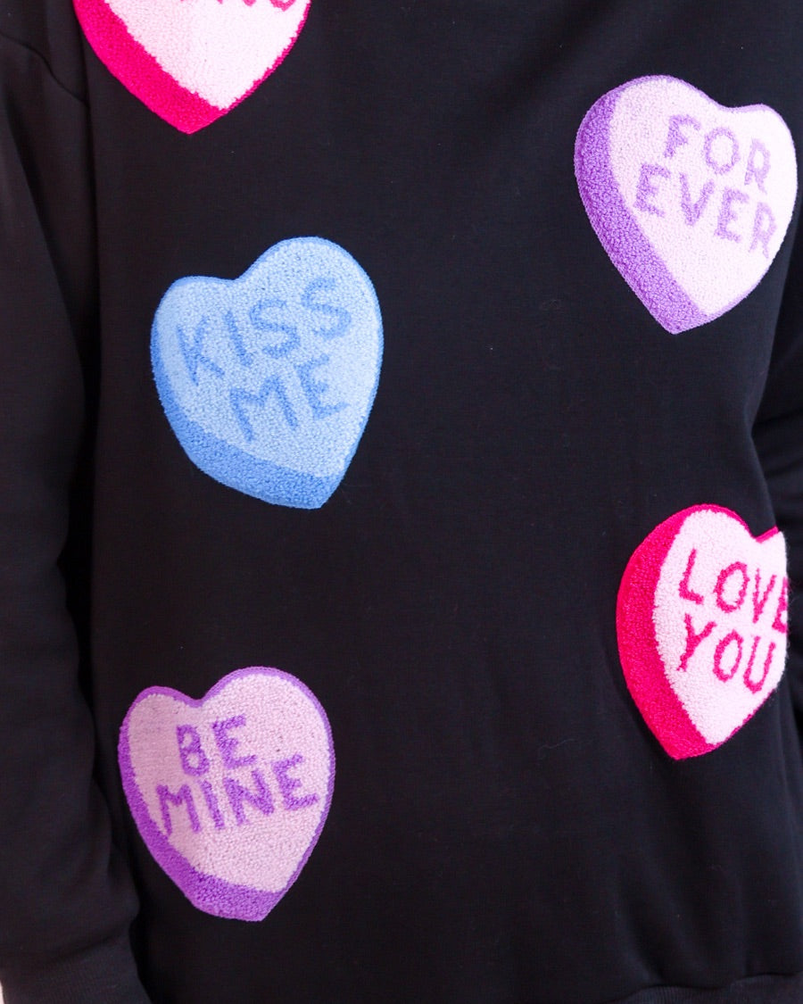 a black long-sleeve sweater made of ultra-soft material adorned with multicolor chenille patch conversation hearts, a relaxed drop-shoulder silhouette, and ribbed trim at the neckline, cuffs, and hemline