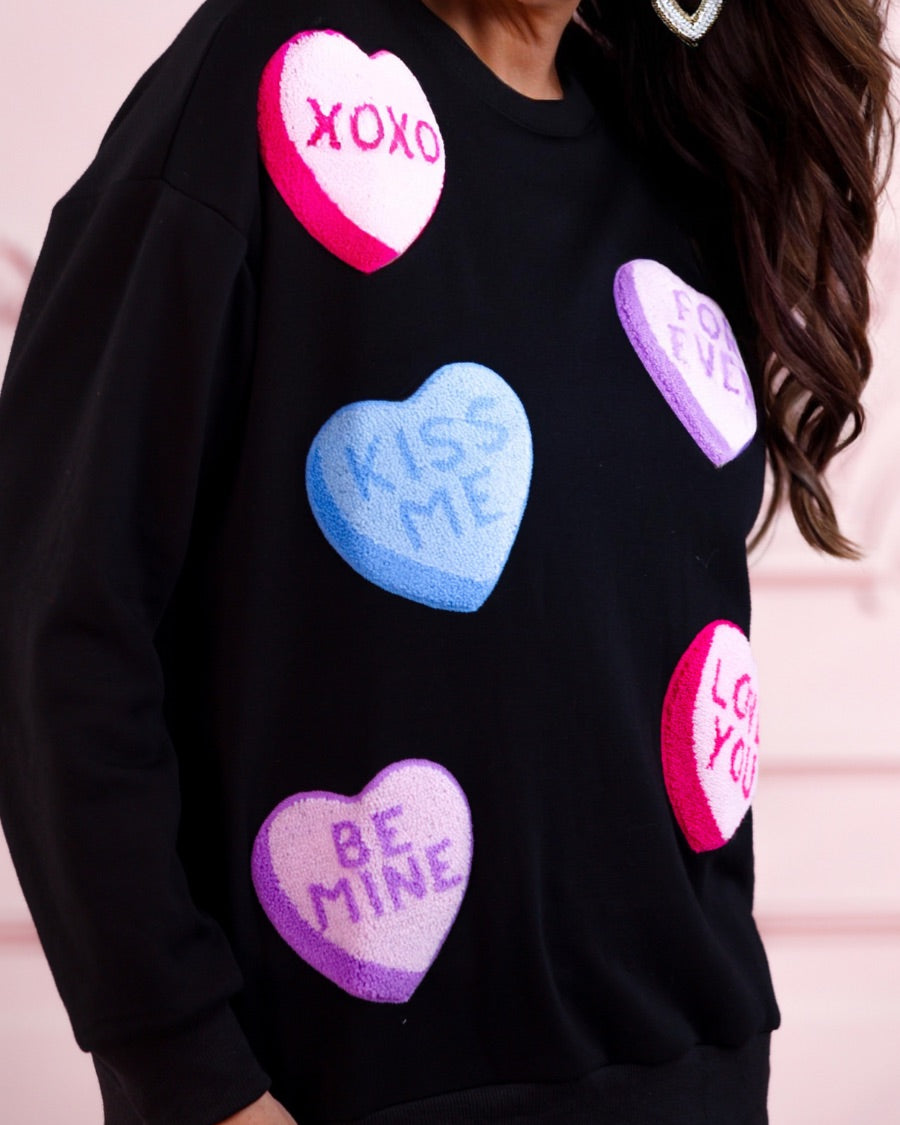 a black long-sleeve sweater made of ultra-soft material adorned with multicolor chenille patch conversation hearts, a relaxed drop-shoulder silhouette, and ribbed trim at the neckline, cuffs, and hemline