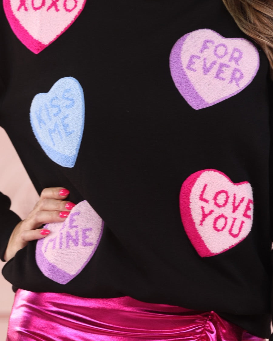 a black long-sleeve sweater made of ultra-soft material adorned with multicolor chenille patch conversation hearts, a relaxed drop-shoulder silhouette, and ribbed trim at the neckline, cuffs, and hemline