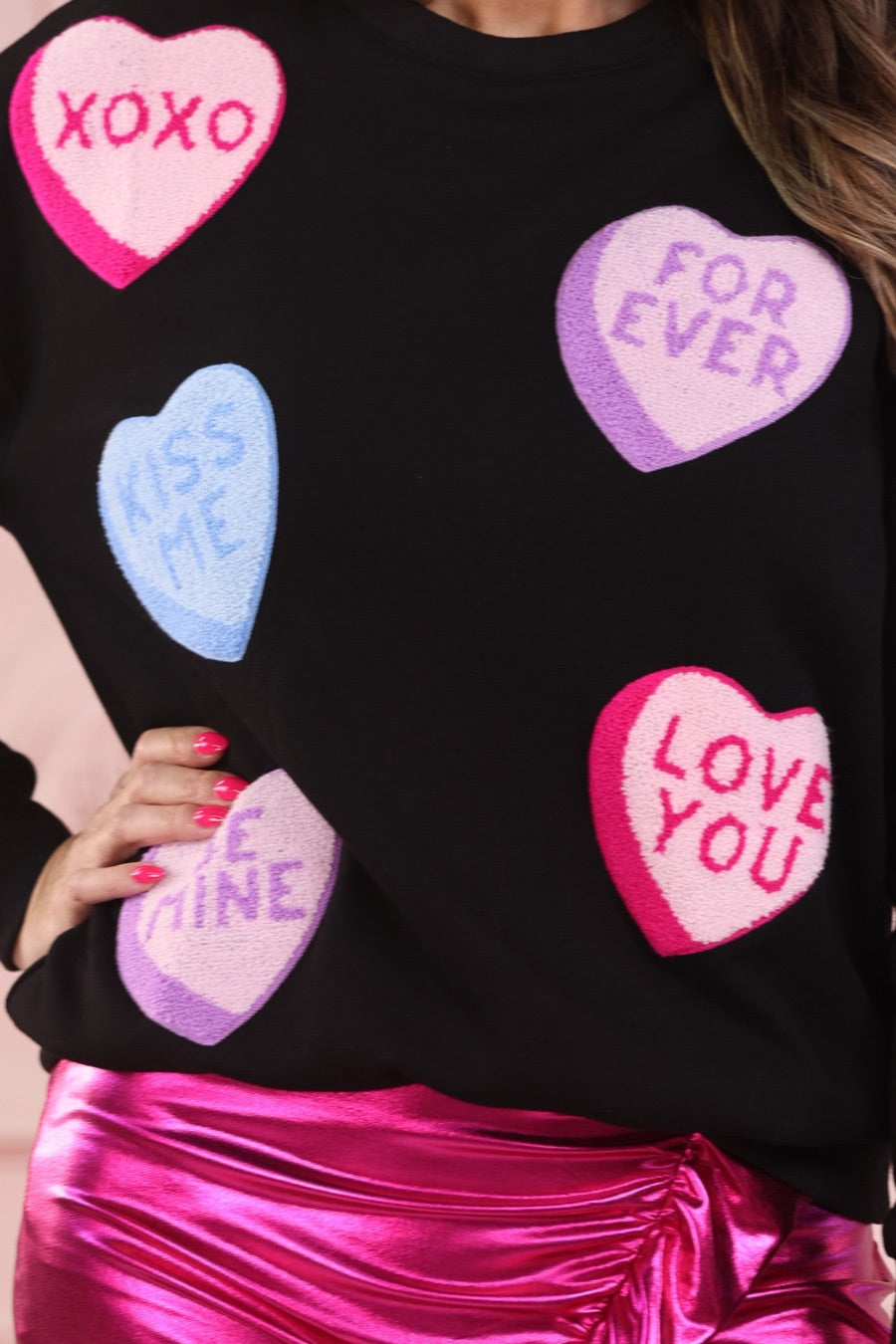 a black long-sleeve sweater made of ultra-soft material adorned with multicolor chenille patch conversation hearts, a relaxed drop-shoulder silhouette, and ribbed trim at the neckline, cuffs, and hemline