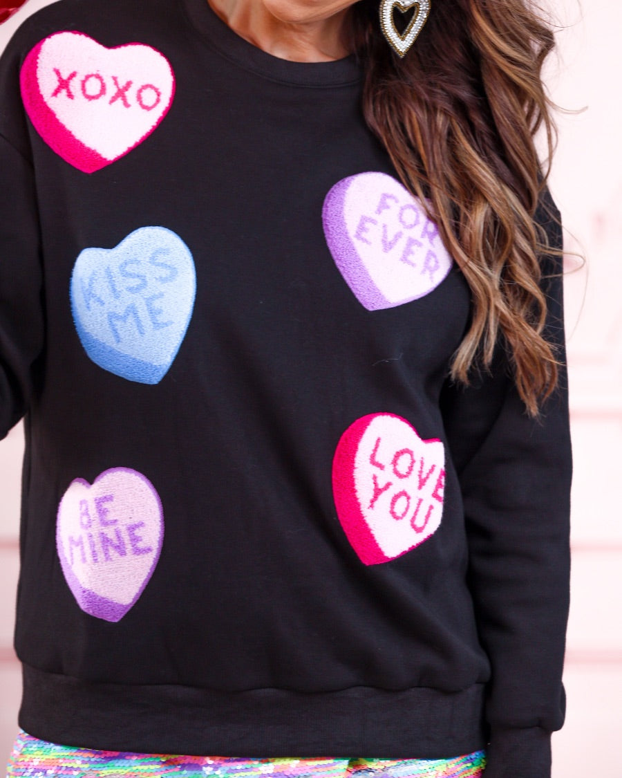 a black long-sleeve sweater made of ultra-soft material adorned with multicolor chenille patch conversation hearts, a relaxed drop-shoulder silhouette, and ribbed trim at the neckline, cuffs, and hemline