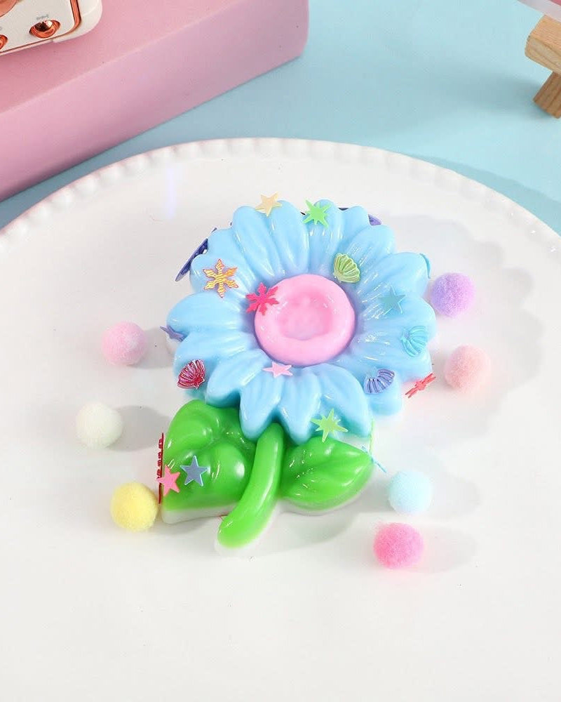 a blue flower-shaped decompression squishy toy with glossy petals, green leaves, mini decorative accents, and pastel pom poms