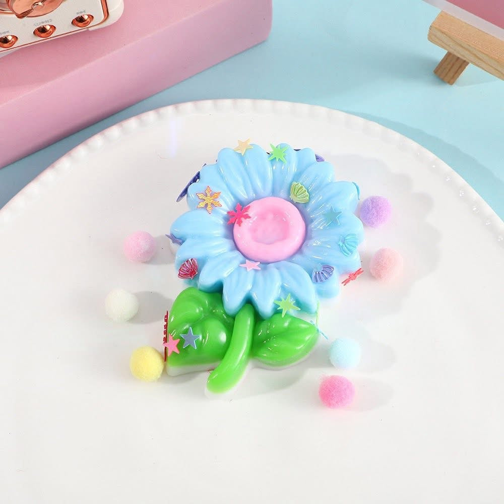a blue flower-shaped decompression squishy toy with glossy petals, green leaves, mini decorative accents, and pastel pom poms