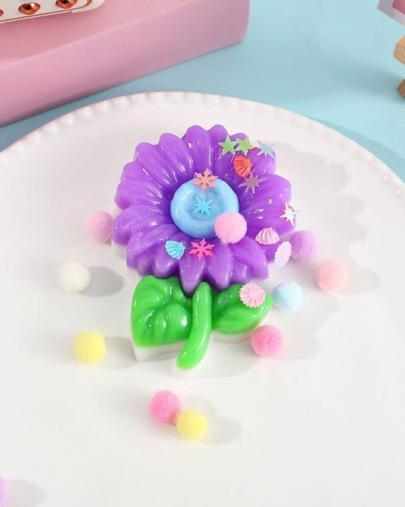 a purple flower-shaped decompression squishy toy with glossy petals, green leaves, mini decorative accents, and pastel pom poms