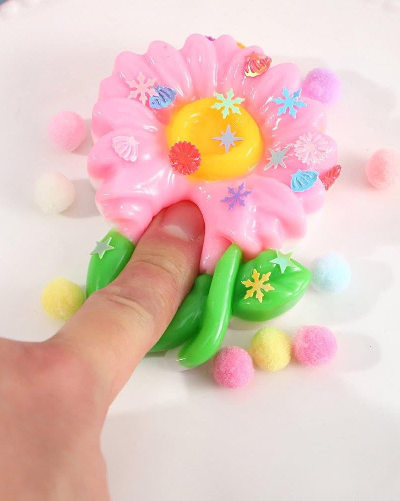 a pink flower-shaped decompression squishy toy with glossy petals, green leaves, mini decorative accents, and pastel pom poms
