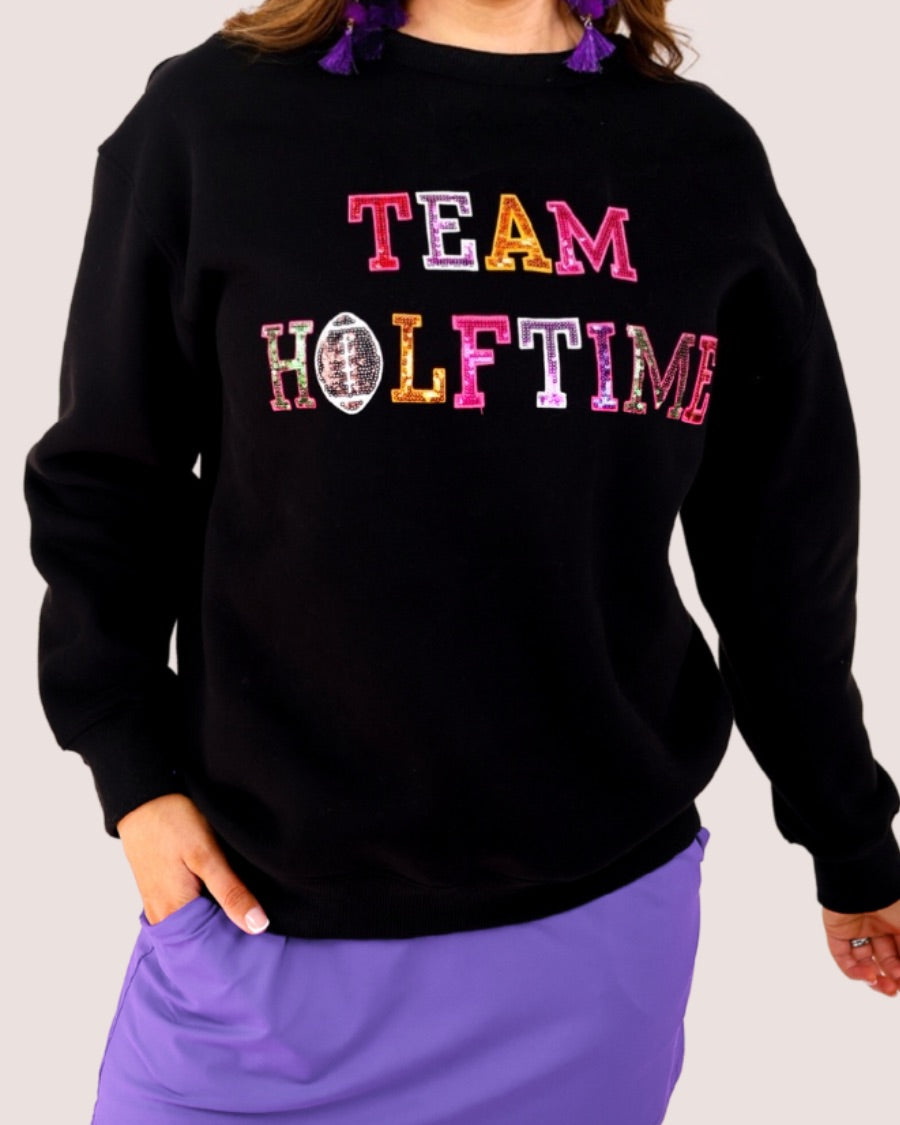 black long-sleeve sweatshirt made from soft mid-weight fabric with a crew neckline, straight hem, and bold multicolored varsity-style chenille patches that spell out 'TEAM HALFTIME' across the front chest