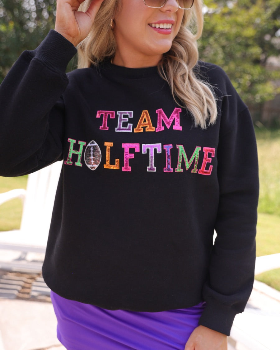 black long-sleeve sweatshirt made from soft mid-weight fabric with a crew neckline, straight hem, and bold multicolored varsity-style chenille patches that spell out 'TEAM HALFTIME' across the front chest